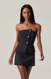 A woman is wearing a strapless, dark-colored, sparkly mini dress with a subtle shimmer. The dress features a vertical row of decorative buttons along the front, adding an elegant touch. The fabric appears textured with a glittering effect, giving the outfit a sophisticated and glamorous look.